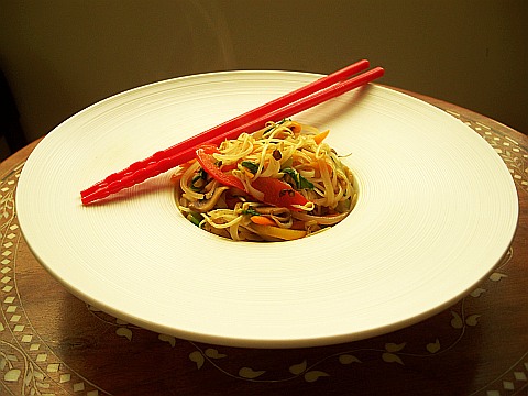 Photo of Pad Kapraw Thai Rice Noodles on a wide-rimmed white presentation plate with red chopsticks